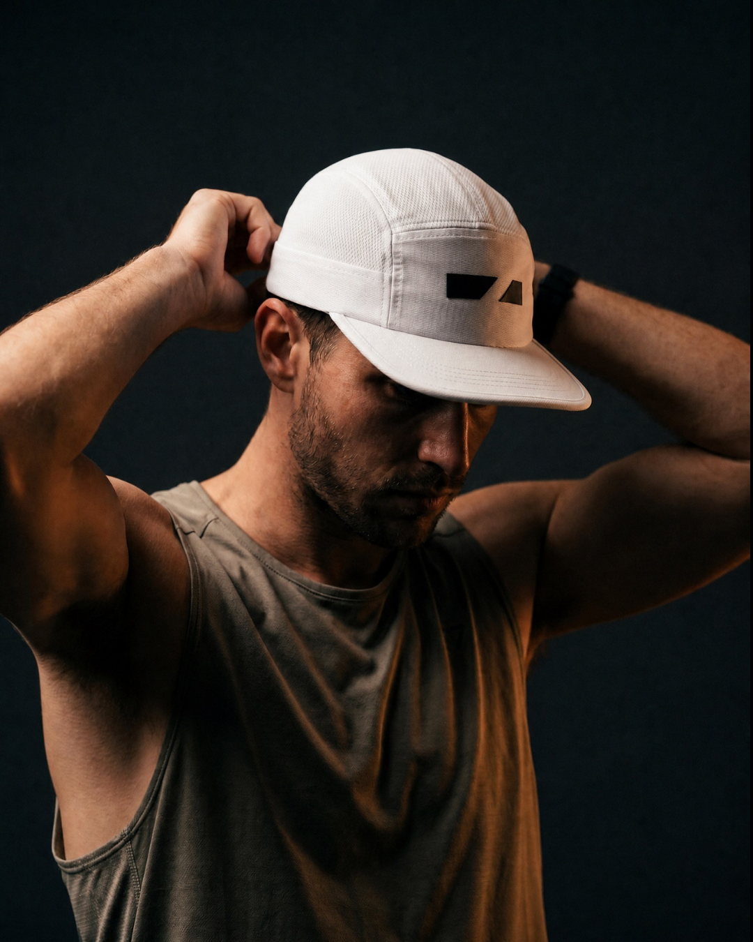 A man wearing a white structured baseball cap with a black logo patch on the front, adjusting the brim with both hands against a dark studio background.