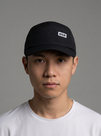 A man wearing a black baseball cap with a white rectangular patch featuring the text 'NSR' on the front, paired with a white t-shirt against a solid gray background.